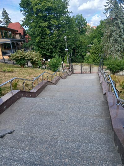 The image shows a wide granite staircase leading down from a modern building. The building, partially visible on the left, has a wooden exterior and large windows, and is topped with a red tiled roof. The staircase is flanked by polished granite walls with stainless steel railings. The steps are made of speckled grey granite. At the bottom of the stairs, there is a black metal gate with an ornate circular design. Beyond the gate, there is a grassy area with some bushes and trees. In the background, more trees and some buildings can be seen on a hillside under a cloudy sky. The overall scene suggests a well-maintained outdoor area adjacent to a building, possibly a hotel or residential complex. The location is identified as Truskavets, Ukraine.