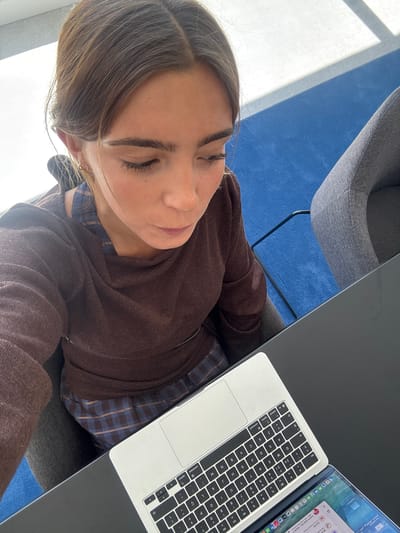 A woman is sitting at a desk in Torres Vedras, Portugal, working on a laptop. The background includes a blue carpet and a grey chair. The woman wears a brown sweatshirt and a blue and brown plaid shirt.