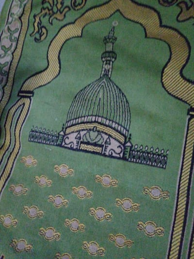 The image displays a green woven prayer mat, positioned flat in the city of Olambe, Nigeria, observed from an elevated, angled perspective. The mat's primary design element is a stylized mosque or sacred building, framed by an arched structure in the upper half. This central motif features a prominent green dome with vertical ridging and a yellow base, topped by a slender minaret-like spire with a finial. Below the dome, a horizontal decorative band incorporates a dark green and yellow fence-like pattern. The arch framing this design is delineated with yellow stripes and black outlines. The lower section of the mat's field is patterned with scattered, repeating oval-shaped motifs in light beige with yellow outlines. The left and right edges of the mat feature vertical borders composed of yellow stripes, black lines, and curved decorative elements on a green background. The textile material exhibits characteristics of a prayer rug, with minor surface variations under ambient lighting. No individuals or active subjects are present within the frame.