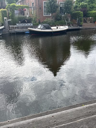 A small, black and white motorboat with a black canopy is moored to a wooden dock on a canal in Katwijk, Netherlands. The boat is dark with a white deck and has a single helm. A blue float is attached to the boat by a rope. The water is dark and reflects the surrounding buildings and trees, creating a rippled, distorted image. The buildings are brick with white-trimmed windows and are surrounded by lush green trees and bushes. A wooden deck with horizontal slats is visible in the foreground.