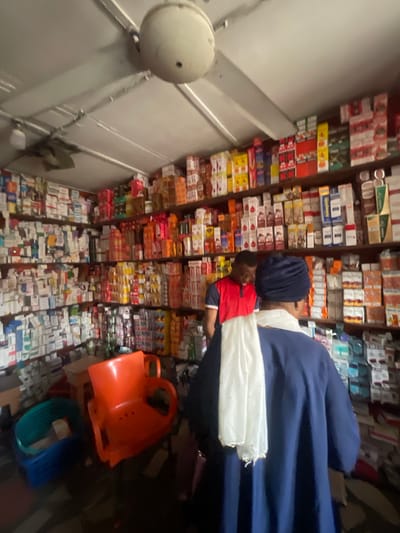 The image shows a bustling pharmacy in Akowonjo, Nigeria, densely stocked with a variety of medications and health products. Shelves line the walls from floor to ceiling, filled with numerous boxes and containers. Two individuals are visible in the frame: a man in a red and blue t-shirt is standing, possibly attending to a customer, while a woman in a blue robe and head covering is facing away from the camera, also possibly a customer or employee. In the foreground, two colorful plastic chairs, one orange and one blue, suggest a waiting area or a place for customers to sit. The lighting is somewhat dim, typical of a small retail space. The overall impression is one of a busy, well-supplied community pharmacy in Nigeria.