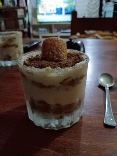 A glass of tiramisu topped with a cookie and dusted with cocoa powder sits on a wooden table. The glass is partially filled with layered creamy, cake, and chocolate components, visible through the glass. There is another glass of dessert on the left, a spoon on the right, and a blurred background showing a TV and other furniture. The scene appears to be in an indoor setting, with the wooden table in the foreground. The coordinates (-34.473, -58.639) place the location in El Talar, Argentina. The scene is well-lit, providing clear visibility of the desserts.