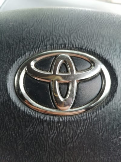 The image shows a close-up of the Toyota logo on a black, textured surface, likely a car's steering wheel. The logo is a silver oval with three interconnected ellipses inside. The black surface has a wavy pattern, characteristic of leather or a similar material used in car interiors. There are no people, actions, or interactions visible in the image. The environment appears to be indoors, possibly within a vehicle. There is no information to place this image in Lesozavodsk, Russia.