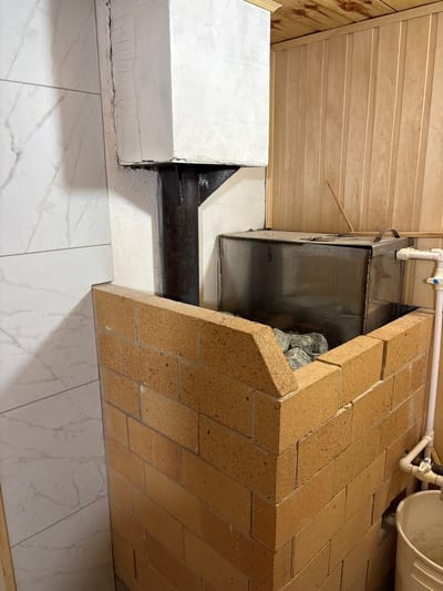 In the city of Ilpanur, Russia, a Finnish sauna is pictured. The focal point is a brick oven with a metal enclosure filled with rocks, likely for heating. To the left, a wall is clad in white tiles with a marbled pattern. To the right and overhead, the wall and ceiling are constructed of light-colored wood planks. A metal pipe protrudes from the oven and extends upwards, surrounded by a white, squared structure. Adjacent to the oven, plumbing fixtures, including a pipe and valve, are visible, along with a white bucket situated on the floor.