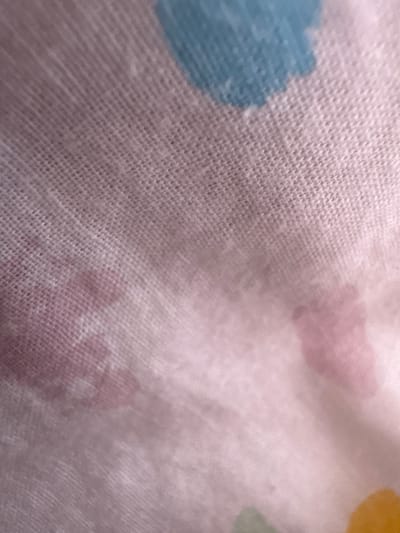 The image displays a close-up, soft-focus view of a light-colored woven textile fabric, likely white or off-white. The fabric exhibits a discernible, fine crisscross weave pattern across its surface. Irregular, diffused splotches of color are present, primarily light blue, light pink, and faint light green/yellow. No individuals, specific objects beyond the fabric, or identifiable actions are visible. The environment is limited to the texture and coloration of the fabric itself. This scene is located in the city of Bukovo, Bulgaria.