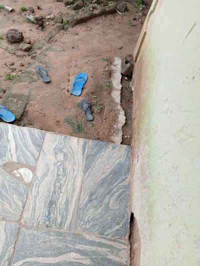 A high-angle shot captures a collection of discarded footwear on the damp, sandy ground beside a light green, textured wall. The scene appears to be located outdoors, potentially in a yard or pathway. Several pairs of flip-flops are scattered across the dirt, including a blue pair near the center and a dark gray pair to the left. The ground is a mix of brown soil, scattered pebbles, and patches of green vegetation. A paved area with large, grey and white marbled tiles is visible in the foreground, leading away from the wall. The wall itself exhibits signs of weathering, with streaks of green and brown discoloration. The overall impression is one of abandonment and neglect. The location is Gindin Dutse, Nigeria.