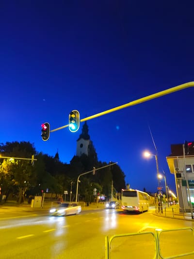 An eye-level, full shot captures a busy city street in Stryków, Poland, at dusk. The sky is a deep, cloudless blue, transitioning into the darkness of night. Traffic lights hang over the road, with one displaying a red light and another showing a green light. A white bus and several cars move along the illuminated street, their headlights cutting through the twilight. Trees line the sidewalks, and a church steeple is visible in the background, adding a touch of architectural interest to the urban landscape. The streetlights cast a warm glow, creating a vibrant atmosphere.