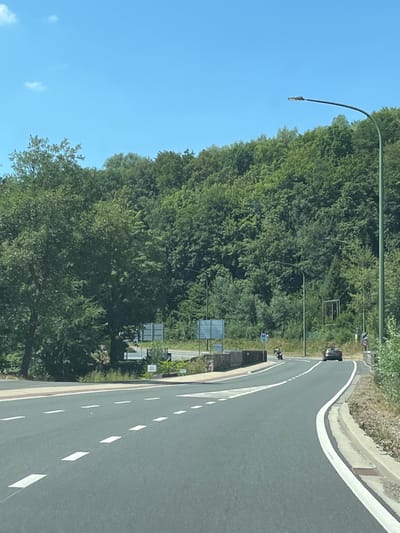 The image depicts a view from a vehicle driving on a paved road in Malmedy, Belgium. The road curves to the right, with white dashed lines marking the lanes. On the left side of the road, there is a sidewalk and a small section of a stone wall. Beyond the wall, a densely wooded hillside rises steeply. A large tree with green foliage is prominent on the left.

In the middle distance, a road passes over a bridge or overpass. A motorcycle is visible on this road, traveling from left to right. Further ahead on the road, a dark-colored car is also traveling in the same direction. Several road signs are present, including what appears to be a speed limit sign with "70" on it.

A tall, green lamppost stands on the right side of the road, extending upwards and curving over the roadway. The sky is clear and blue with a few small white clouds. The overall impression is a sunny day in a semi-rural or suburban area with significant tree cover.