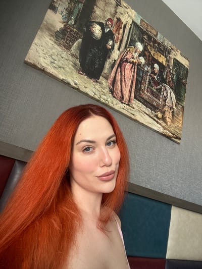 A medium shot photo captures a woman with vibrant, long, orange hair posing in front of a textured grey wall adorned with a painting. The painting depicts a marketplace scene with figures in traditional clothing. The woman is positioned in the foreground, with her face partially visible as she looks towards the camera. Her lips are slightly parted, and she is wearing a light-colored garment. The background features a headboard with distinct color blocks of dark blue, light beige, and dark red. The scene is likely indoors, possibly in a hotel room or residential space, in Avcılar, Turkey.