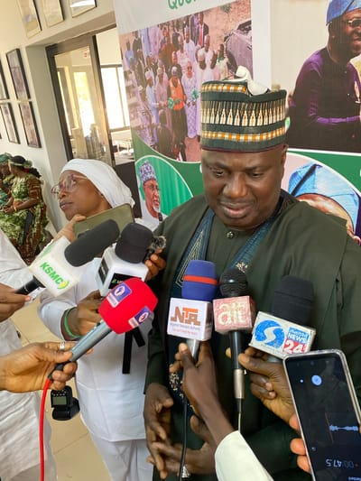 An indoor scene captures a media engagement in Kwarau, Nigeria, featuring a man addressing a group of reporters. The central figure, dressed in an olive-green traditional tunic and a patterned cap, is speaking into multiple microphones. His expression is calm, with a slight smile.
Numerous hands, belonging to various media personnel, are visible extending microphones and recording devices towards the speaker. Prominently displayed microphone logos include "KSMC," "CHANNELS" (on a red microphone), "silverbird NEWS 24," and "NTA" (Nigerian Television Authority), the latter bearing the tagline "You can't beat the reach." One individual is seen holding a smartphone actively recording audio, with a timer showing "00:47.5" and a pause icon on its screen. Another person in a white head covering and glasses is also holding a microphone, suggesting their involvement as a reporter or participant.
In the background, a large green and white banner is partially visible, featuring blurred images of people, some wearing traditional attire, and partial text including "QU." Below the banner, framed pictures adorn a light-colored wall, and a window reflecting bright light is visible further back. The overall setting appears to be a formal press conference or interview session within a building, likely a media center or public office. The artificial lighting or bright window light suggests the event could be taking place during daytime.