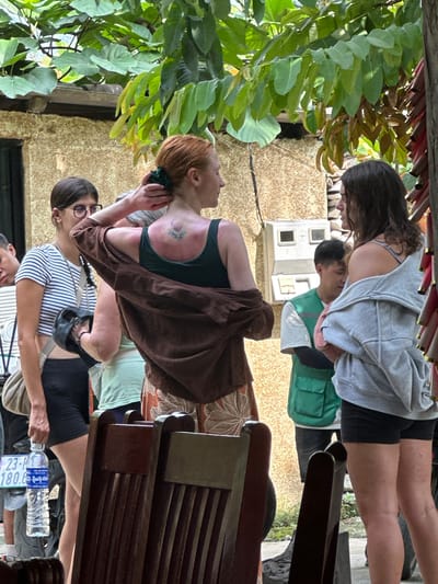 The image, taken in Du Già Commune, Vietnam, captures a bright daytime scene of several individuals gathered in what appears to be an outdoor cafe or seating area. The overall setting is casual, with lush green foliage visible on the left side, and a textured, light brown building wall forming the backdrop.

In the foreground, on the left, a person with dark hair and glasses wears a striped short-sleeved top and black shorts. They hold a clear plastic water bottle with a blue "Country Mile" label in their right hand. Partially visible behind them is a Vietnamese license plate displaying "23-H1 180.0", where "23" denotes Hà Giang province, confirming the location context.

In the midground, a person with red hair is prominent. They are wearing a dark tank top and a brown shawl draped over their shoulders. This individual appears to be either talking on a phone or wiping their face with a small dark cloth held to their ear. A floral tattoo is visible on their left shoulder.

Further in the background, two more individuals are present. One has short dark hair and wears a green vest over a light-colored shirt. Behind them, another person with long dark hair is seen wearing a grey hooded sweatshirt and black shorts. All subjects appear relaxed and engaged in their own activities, possibly socializing or resting. Elements of dark wooden furniture, likely chairs or benches, are visible on the right side of the frame. A white electrical box or utility panel is mounted on the wall in the distant background.