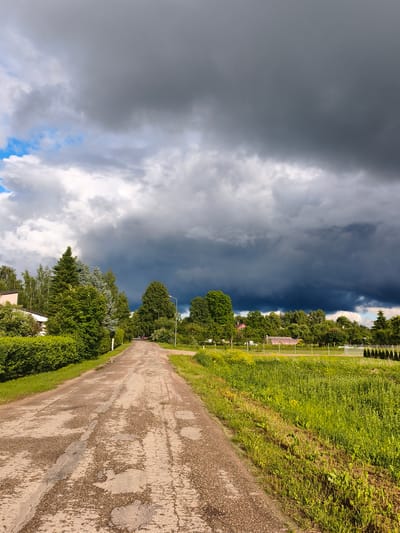 The image captures a rural scene in Brodi, Latvia, under a dramatic sky. A paved, weathered road stretches into the distance, flanked by lush green grass and trees on either side. On the left, a neat hedge lines the road, with houses and more trees visible beyond. To the right, a field of tall green grass with scattered yellow wildflowers dominates the landscape, leading towards more trees and a few structures, possibly a greenhouse or sheds, in the background. The sky is filled with dark, heavy storm clouds, creating a stark contrast with patches of bright blue sky and white clouds visible to the upper left. A single streetlamp stands on the right side of the road. The overall impression is one of impending weather in a quiet, green setting.