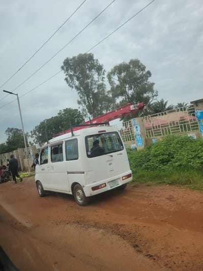 The image shows a white utility van parked on a dirt road. A red ladder is extended from the side of the van, suggesting it is being used for some sort of work. The van appears to be a Daihatsu Hijet. The scene is set in Bokkos, Nigeria.
In the background, there is a building with a metal fence and some greenery. There are also several posters on the building, some of which have images of people and text, likely political campaign materials. The sky is overcast, indicating cloudy weather.
There are a few people visible near the van and the building. One person is standing near the front of the van, and others are gathered near the building. It's unclear what specific activity is taking place, but the presence of the ladder and the van suggests some form of repair, maintenance, or construction work. The lighting suggests it is daytime, but the cloudy sky makes it difficult to determine the exact time. The overall mood of the image is neutral, with no obvious signs of distress or excitement.