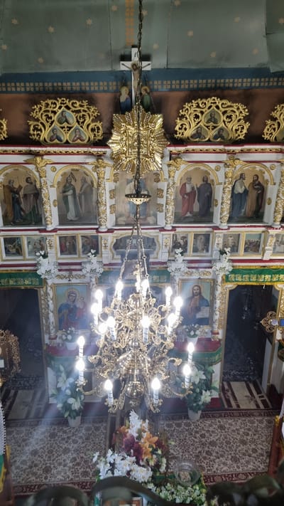 The image shows the interior of an Orthodox church, likely in the city of Vyshka, Ukraine, viewed from above. A large, ornate golden chandelier hangs prominently in the center, illuminated by multiple light bulbs. The walls are decorated with religious icons and gilded ornamental carvings. In the background, a cross with a crucifix is visible, along with more icons depicting religious figures. The floor is covered with a patterned carpet. Floral arrangements are placed on the floor and near the altar area. The overall impression is of a richly decorated and solemn religious space.