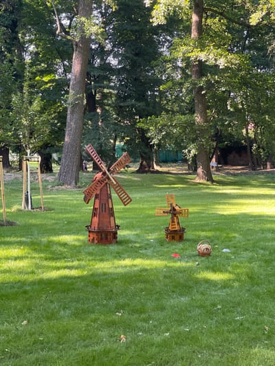 The image depicts a grassy park area in Stupava, Slovakia, dominated by lush green grass and surrounded by mature trees. In the foreground, two decorative wooden windmills are placed on the lawn. The larger windmill, on the left, is a detailed replica with four blades and a multi-story structure. To its right and slightly further back, a smaller, similar windmill stands. A small woven basket containing what appears to be rope is situated between the two windmills. In the background, more trees are visible, and hints of human figures can be seen among them. The lighting suggests it is daytime, with sunlight filtering through the leaves, casting shadows on the grass.