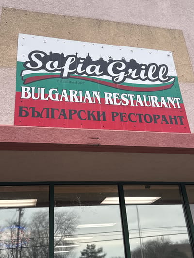 The image shows the exterior of the Sofia Grill restaurant in Des Plaines, Illinois, USA. The sign displays "Sofia Grill" in stylized lettering above an outline of a cityscape. Below this is "Bulgarian Restaurant" in English and Bulgarian. The sign is mounted on a building. Adjacent to the sign is a window reflecting the outside environment.
