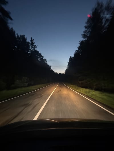 The image depicts a view from the driver's perspective, looking forward along a dark asphalt road through a forest. The sky above is a deep twilight blue, with a lighter gradient near the horizon. The road has a white line marking the center and white lines on the edges. Headlights from the vehicle illuminate the road ahead, casting a warm glow. Tall, dark pine trees line both sides of the road, creating a sense of enclosure. The overall impression is that of driving on a rural road at dusk or early night. The location, Балаки, Russia, is a rural area, consistent with the visual cues of a forest road. There are no people or other vehicles visible in the image. The image is a photograph taken from within a moving vehicle.