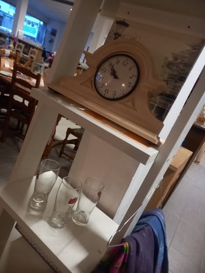 A tilted, high-angle shot shows a shelf with decorative elements. An off-white clock is on the upper-left. A stack of three empty glass bottles, each with a small red mark, are on the white shelf on the right. A blue and purple fabric is folded nearby. In the background, there are tables, chairs, and store displays.