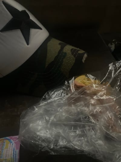 The image shows a close-up of a white cap with a black star logo, placed on a surface that appears to be a table. Adjacent to the cap is a plastic bag containing what looks like fruit, possibly a peach or apricot, sliced and partially visible. The background is dark and out of focus, with some texture suggesting a wooden surface. There is no information in the image to indicate the location in the city of Tavo, Nigeria.