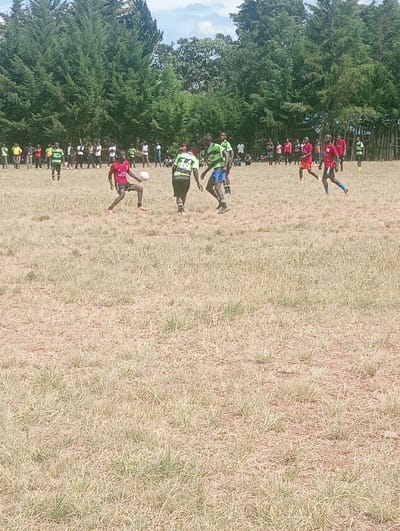 An outdoor soccer match is underway on a dry, light brown grassy field in Lwandeti ward, Kenya, under a partially cloudy sky. In the foreground, a male player wearing a red shirt with the number 10 and dark shorts actively dribbles a white soccer ball with his right foot. To his right, a second male player in a green and black striped shirt with the number 24 and dark shorts is positioned facing away. A third male player, also in a green and black striped shirt and blue shorts, is to the right of the player with the ball, facing left. Further to the right, a fourth male player in a red shirt and blue shorts is in motion towards the left. Additional players, dressed in red or green and black striped shirts, are visible further across the field. A large group of spectators in varied casual attire stands and sits along the perimeter of the field in the background, observing the game. A dense line of tall, dark green coniferous trees forms the backdrop of the scene, with lighter green foliage interspersed. The ground is characterized by sparse, dry, light brown grass with some small patches of green.