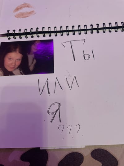 A photograph shows an open spiral-bound notebook on a surface that appears to be camouflage fabric. On the left page of the notebook, there is a lipstick imprint. To the right of the lipstick imprint, there are three lines of handwritten Russian text. The first line reads "Ты" (Ty, meaning "You"). The second line reads "или" (ili, meaning "or"). The third line reads "я" (ya, meaning "I"), followed by three question marks. There is also a small, blurry photograph inserted into the notebook, showing a young woman with dark hair. The notebook is positioned on a piece of camouflage patterned fabric. No location in Vaganovo, Russia is identifiable in this image.