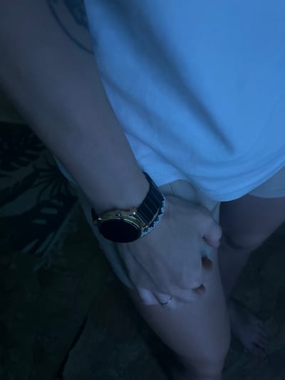The image shows a close-up view of a person's arms and legs. The person is wearing a white t-shirt and light-colored shorts. On their left wrist, they are wearing a smartwatch with a black band and a gold-colored bezel. A bracelet with a pattern of white flowers and black beads is also visible on the same wrist. The person's hands are clasped together, and a thin silver ring is visible on the ring finger of their right hand. The background is dark and appears to be a floor with a textured pattern, possibly a rug or carpet. The lighting suggests an indoor setting, with a blueish tint. There is no indication of the specific location within the city of Ostróda, Poland.