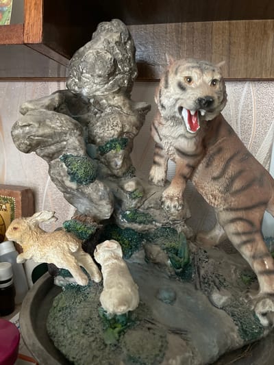 This is a close-up, tilted shot of a decorative figurine featuring a tiger and two rabbits on a rocky, naturalistic base. The tiger, depicted mid-pounce with its mouth open and tongue visible, appears to be lunging towards the rabbits. The rabbits, rendered in a lighter, creamy color, are positioned in front of the tiger. One rabbit is curled up, while the other is reaching out. The base is designed to resemble a rocky terrain with patches of green foliage, suggesting a woodland or jungle setting. The lighting appears to be artificial and is consistent throughout the image. There are no visible time of day or weather cues. The context provided indicates the location as Izborsk, Russia, though there are no visual elements within the image that specifically point to this location. There is no visible text in the image.