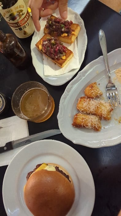 An overhead view captures a dining table set with various food and beverage items. A person's light-skinned hand extends into the upper-left of the frame, positioned above a white rectangular plate holding two pieces of toasted bread topped with a dark, finely chopped mixture, likely beef tartare, featuring small yellow dollops and green specks. Next to this, a bottle of Kirin Ichiban Premium Beer is visible, along with a dark brown glass bottle.
To the right, an irregular-shaped white plate contains three golden-brown fried items, likely potato croquettes, generously topped with grated white cheese; a silver fork rests partially on this plate, indicating prior consumption. A glass tumbler containing amber-colored liquid, presumed to be beer, sits on a coaster below the tartare plate. A serrated knife with a black handle is positioned on a white napkin below the beer glass. At the bottom of the frame, a round white plate holds a hamburger with a brioche bun, a visible meat patty, melted yellow cheese, and a white sauce. The table surface is black.
The specific location within the city of Modiin-Maccabim-Reut, Israel, is not discernible from the visual information provided in the image.