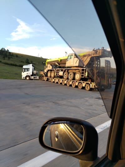 A side view of a large crane truck is captured through a car window on a sunny day. The truck is parked on an asphalt surface, possibly a highway or rest stop, with a grassy hill and trees in the background. The crane truck itself is a complex piece of machinery, with a long boom and multiple wheels. The sun is setting, casting a warm glow on the scene and reflecting in the car's side mirror. There are no people visible in the image. The image appears to be taken in the city of Otarsheni, Georgia, based on the provided context.