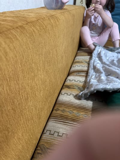 The media file depicts a close-up, low-angle shot of a child lying on a couch in what appears to be a living room. The child, wearing pink pajamas, is propped up on their elbows and eating. A patterned blanket is draped over them and the couch. The couch is upholstered in a textured, mustard-yellow fabric, with a contrasting patterned cushion or bolster behind the child. A clear plastic bottle, possibly a sippy cup, is visible on the left side of the frame, near the child's feet. The lighting suggests an indoor setting, likely during the day. The scene conveys a sense of casual domestic life, possibly a moment of relaxation or snack time. No visible text is present in the image. The location context provided is Shamalgan, Kazakhstan.