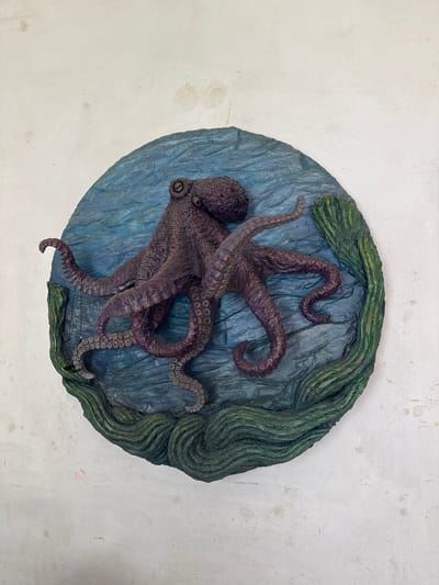 A circular wall plaque depicts a dark purple octopus with eight tentacles spread out on a textured blue background, designed to resemble water. Green, wavy seaweed forms the bottom edge of the plaque. The plaque is affixed to a textured, off-white wall with visible imperfections, including stains and scuffs. The scene is indoors, likely a studio or workshop, and there is no indication of the city of Kolomyia, Ukraine.