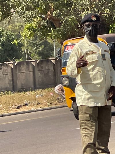 This image depicts a sunlit street scene in the city of Basawa, Nigeria. In the foreground, an adult male with dark complexion stands, facing slightly right. He is dressed in a black beret bearing a prominent circular emblem, a black face mask covering his nose and mouth, a light-colored (cream or khaki) long-sleeved shirt or jacket with multiple pockets, a patch labeled "D490 MO" on his right chest pocket, and a green rectangular insignia on his left chest. He wears olive-green cargo trousers. His right hand is raised to chest height, with a silver-toned ring on his fourth finger, appearing to make a gesture.

Behind and to the left of the man, a yellow motorized rickshaw (keke Napep) is partially visible, showing its front right headlight and part of the cab with red lettering, possibly "Npe." A person in a white and brown striped garment is indistinctly seen near or inside the rickshaw.

The immediate environment consists of a paved road with a faint yellow line. To the left of the road, a strip of dry, light-brown grass is littered with various refuse. Beyond this, a concrete wall, constructed from rectangular panels with angular, possibly decorative, protrusions along its top edge, spans the mid-ground. Dense green trees and foliage are visible above and behind the wall, under clear skies.
