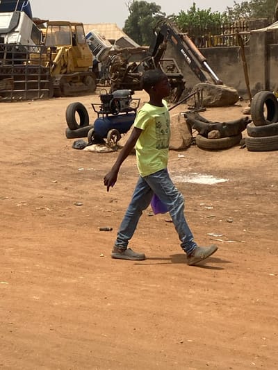A young male individual with dark skin and short dark hair is depicted walking from right to left across a reddish-brown dirt surface. He is wearing a light lime green short-sleeved t-shirt with a faded graphic print on the front, light blue denim jeans, and gray-brown slip-on shoes. A small, folded purple object is held in his left hand. His head is turned slightly towards the right, while his body is oriented to the left. The background features an outdoor industrial or scrap yard environment under daylight. Visible machinery includes a large yellow tracked bulldozer on the left, a blue wheeled air compressor with an engine in the center, and a black hydraulic excavator arm labeled "ATLAS 90.1" on the right. Numerous black rubber tires and large rocks are scattered across the uneven ground. A white vehicle frame is partially visible on the far left. Distant background elements include simple buildings and sparse trees. Patches of lighter-colored residue are present on the dirt ground. The scene is located within the city of 0801712591, Nigeria.