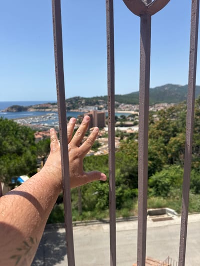 A close-up of a person's hand reaching out through a metal fence reveals a sun-drenched landscape. The hand, glistening with sweat, is positioned in the foreground, partially obscured by the rusty metal bars. Beyond the fence, a coastal city unfolds under a clear blue sky. Buildings with terracotta roofs dot the hillside, leading down to a marina filled with boats. The sea stretches to the horizon, meeting a distant, tree-covered mountain range. The scene evokes a sense of longing, with the hand reaching towards the freedom of the open view.