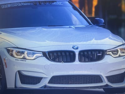 The image is a close-up, low-angle shot of the front of a white BMW M3. The car is white with black accents, including the grille and side mirrors. A "RECHARGE RALLY" sticker is visible across the top of the windshield. The car is parked on a street, and the background appears to be slightly blurred, suggesting it's outdoors. There are no people visible in the image. The car is clean and appears to be in good condition. The lighting suggests it is daytime. The specific location in the city of Pereiaslavske, Ukraine, cannot be determined from the image alone.