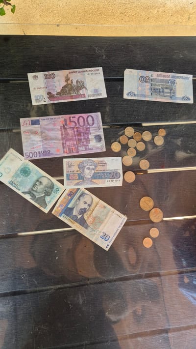 In the city of Butovo, Bulgaria, on a dark wooden table, are various currencies. A 100 Russian ruble note and a 50 Russian ruble note are placed at the top of the table. Below these are a 500 Euro note, a 50 Norwegian krone note, a 20 Bulgarian lev note, and another 20 Bulgarian lev note. Scattered around the notes are several copper-colored coins. The scene is lit by natural light, casting faint shadows.