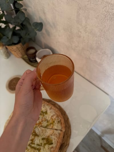 A hand holds a ribbed, amber-colored glass mug filled with a light brown liquid. The mug is positioned over a round pizza on a woven mat. In the background, a green plant in a basket is visible, along with a textured wall. The scene appears to be indoors, likely a kitchen or dining area. No specific location in Semiluki, Russia, can be determined from the image.