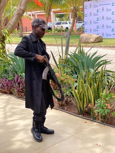 A dark-skinned man, appearing to be security personnel, stands prominently in an outdoor setting, holding an AK-style assault rifle with a silver magazine. He is dressed in a dark, possibly black, long-sleeved shirt or jacket, dark trousers, and black combat boots, topped with a camouflage-patterned baseball cap.
The scene features lush tropical vegetation, including palm trees with orange-red blossoms, agave-like plants, and variegated snake plants (Sansevieria trifasciata) with green and yellow stripes. The man stands on a light-colored tiled path, which borders a garden bed.
In the background, a large white banner is visible, indicating a formal event. Several vehicles, including a white Toyota pickup truck, are parked behind the banner. A large, natural rock is situated near the base of the banner. The bright lighting suggests it is daytime. The overall impression is an event taking place in a warm, possibly tropical, outdoor environment in Kwarau, Nigeria, with a visible security presence.
Visible text on the banner includes "Mid-Term Review," "ACRESAL," "THE WORLD BANK," and "NIGERIA" (accompanied by the Nigerian coat of arms), repeated multiple times. The word "TOYOTA" is also partially visible on one of the vehicles.