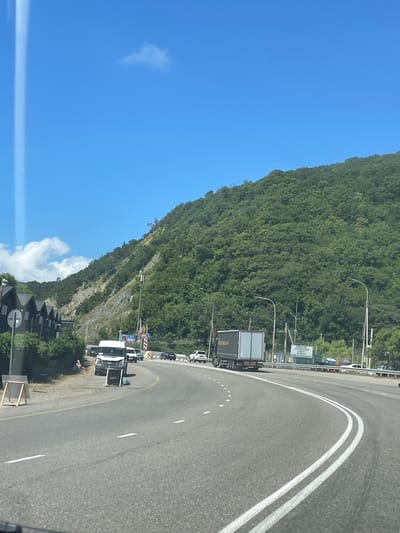 A bright, sunny day is depicted in Lermontovo, Russia, with a clear blue sky and scattered white clouds. A winding asphalt road dominates the foreground, with a truck in the right lane and several cars in the left lane. A steep, densely wooded mountain rises on the right side of the road, its green foliage contrasting with the rocky, barren slope on the left.
Along the left side of the road, a row of dark, modern houses with pointed roofs is visible. A person is sitting on the side of the road near a white van, with a wooden easel and a small table placed nearby. Road signs, including a traffic light and directional signs, are present along the roadside. The scene suggests a busy travel route through a scenic, mountainous coastal area.