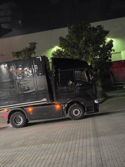 A black semi-trailer truck is parked at night on a paved lot in front of a light-colored building with green fluorescent lighting. The truck has a dark trailer with a light-colored airplane graphic on its side. The truck's cab is black and has the text "TGX 18.480" on the side. The parking lot is made of grey interlocking pavers. Trees with dark green foliage are visible behind the truck and trailer. The sky is dark and appears to be night. No people are visible in the image.