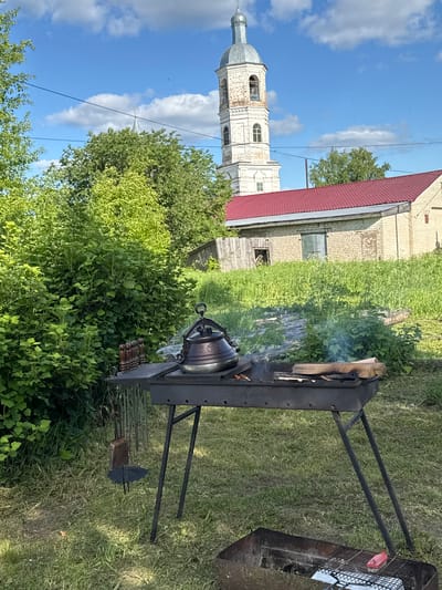 The image captures an outdoor scene set in what appears to be a rural or semi-rural environment in Луптюг, Russia, under a clear blue sky with scattered white clouds. The ground is covered with green grass and lush bushes, suggesting a warm, sunny day.
In the mid-ground and foreground, an outdoor cooking setup is visible. A dark metal brazier or grill stand holds a cast-iron kettle, possibly a kazan or Dutch oven, placed directly over burning wood and coals. Smoke gently rises from the fire, indicating active cooking. A large piece of wood is still burning on the left side of the brazier. Below the kettle, several metal skewers with wooden handles are arranged in a holder, suggesting preparations for grilling meat or vegetables. To the right of the kettle, another section of the grill features a metal grate, on which a red object (possibly a piece of food or a container) rests next to a white cloth or paper. Further right on the ground, a metal tool, resembling a shovel or ash rake, is present.
In the background, a striking architectural element dominates the left side of the frame: a historic, light-colored bell tower or church steeple with a domed, silvery roof. Its weathered facade suggests age and historical significance. Behind the bell tower and extending towards the right, a building with a bright red gabled roof and light-colored brick walls is partially visible. The overall scene depicts a peaceful moment of outdoor cooking, likely a leisure activity, with a backdrop of traditional Russian architecture and nature. There is no visible text in the image.