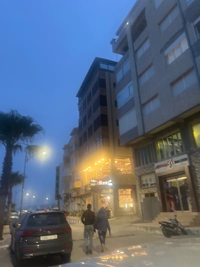 The image depicts a street scene in Fnideq, Morocco, taken during the evening. Tall buildings line the street, some with visible storefronts. Pedestrians are walking on the sidewalk. A parked car is visible in the foreground. The sky is a dark blue, suggesting it is either dusk or dawn. Palm trees are visible in the lower part of the frame, adding to the locale. Streetlights illuminate the scene.