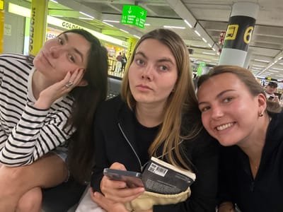 Three adult females are seated in a public waiting area within Palma de Mallorca Airport (PMI) in Son Sant Joan, Spain. The woman on the left, with long dark hair, wears a black and white striped long-sleeved shirt, has red nail polish, and poses with her right hand resting near her face, displaying a ring on her right ring finger. The central woman, with light brown hair, holds a smartphone in her left hand and a cream-colored pouch containing a black-covered book, presenting a neutral expression towards the camera. The woman on the right, with brown hair, smiles broadly at the camera. The immediate background shows a "GOLDCAR" sign, an overhead green sign indicating "Lavabos Toilets Aseos" with directional arrows, and a support pillar marked with a bus icon and the number "6". The environment is brightly lit and features a structured ceiling, consistent with an airport terminal. Other indistinct figures are visible in the distant background.
