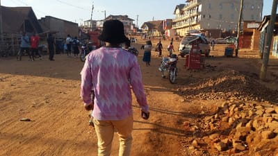 The image shows a street scene in Kayna, Democratic Republic of the Congo. The photo is taken from a low angle, looking towards a man wearing a pink and white patterned shirt and a black hat. He is walking toward the camera, and he is the main subject of the photograph. Behind him, to the right, a motorcycle is parked in the middle of the street. To the left, several other people are walking, and there are several buildings in the background. The road surface is dirt, and there are piles of rocks on the sides of the road. The scene is typical of a busy street in a developing country. There are a few businesses visible in the background, including a car dealership and a small store. The scene is lit by the sun, and the sky is clear.
