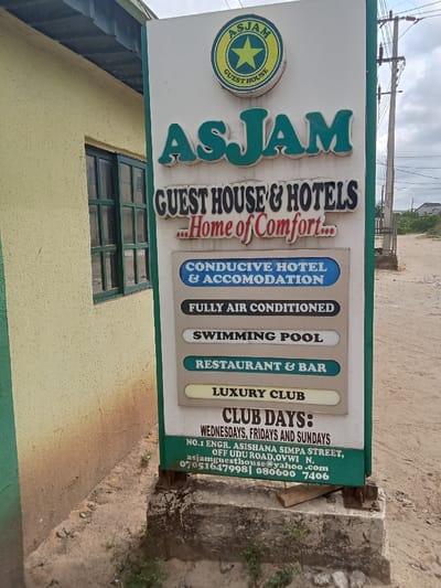 The image shows a sign for a guest house and hotel called "ASJAM" located at No. 1 Engr. Asishana Simpa Street, off Udu Road, Ovwvi N. in the city of Ovwian, Nigeria. The sign is white with green lettering and features a star in a circle with the words "Guest House" around it. The sign also lists the following amenities: Conducive Hotel & Accommodation, Fully Air Conditioned, Swimming Pool, Restaurant & Bar, and Luxury Club. The sign also states that the hotel is open on Wednesdays, Fridays, and Sundays. The sign is positioned in front of a yellow building. The ground surrounding the sign is covered in sand.