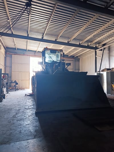 This image depicts the interior of a workshop or storage facility, likely in Makariv, Ukraine. The dominant subject is a large, yellow front-end loader parked prominently in the center of the frame. Its massive bucket is lowered, casting a significant shadow on the concrete floor. The loader appears to be stationary, with no activity taking place.
The setting is a large, well-lit industrial space. The ceiling is constructed with corrugated metal panels supported by dark metal beams, and a chain hoist system is visible hanging from the rafters. Natural light streams in from large, open sliding doors at the far end of the building, illuminating the space and creating a contrast between the brightly lit entrance and the shadowed interior. The walls are also clad in corrugated metal, with some sections appearing to be white or light gray.
To the left of the loader, a workbench with tools and equipment is partially visible, suggesting that the space is actively used for maintenance or other work. There are various metal shelves and storage units scattered throughout the area. The floor is concrete, showing some wear and tear consistent with an industrial environment.
The time of day is suggested by the strong natural light entering the doors, indicating it is likely daytime. There are no people visible in the image. The overall mood is one of stillness and functionality, hinting at a workspace that is either at rest or preparing for action. No legible text is discernible in the image.