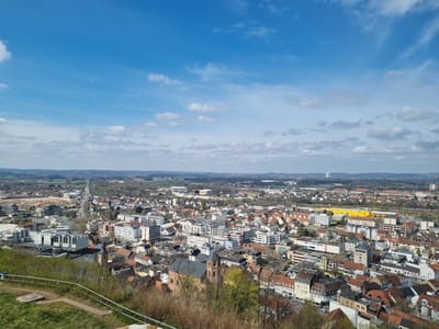 The image presents an elevated view of Homburg, Germany, captured on a bright day with a predominantly blue sky scattered with white clouds. The cityscape is a mix of residential and commercial buildings, with various architectural styles and sizes. Dominating the foreground are older structures with red-tiled roofs. Interspersed are modern, white-facade buildings with flat roofs. 

A church with a prominent bell tower stands centrally within the frame. Distant landscapes show expanses of trees and fields. In the far background, a tall, industrial chimney can be seen. The photo is taken from an elevated vantage point, with a glimpse of greenery and a metal safety railing in the immediate foreground. No people are directly visible in the scene.