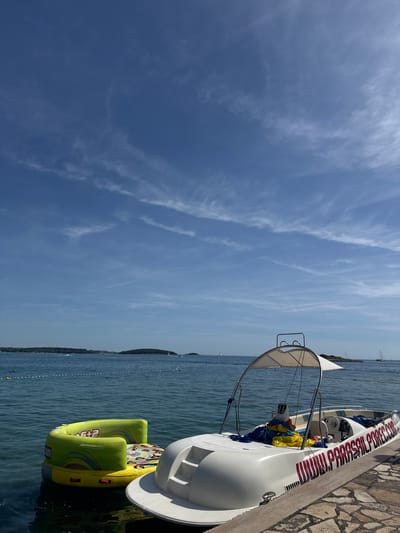 The image depicts a boat docked at a stone pier in Općina Funtana, Croatia. The boat is white with red lettering. Green and yellow inflatable tubes are attached to the side of the boat. The water is calm and there are a few small boats in the distance. The sky is blue with wispy clouds.