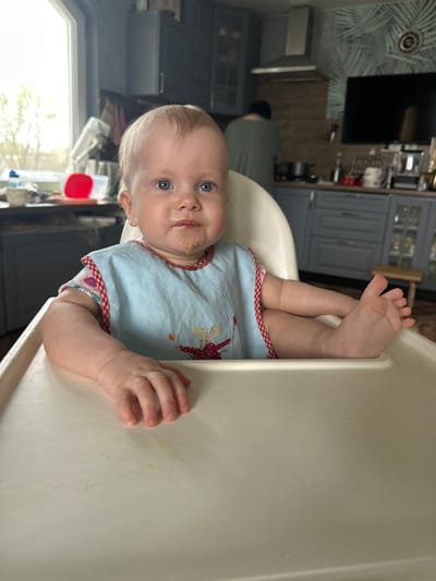 A baby with blue eyes and fair hair is seated in a white high chair, wearing a bib with a red and white checkered trim and a light blue body. The baby's feet are raised, toes pointing upwards. There appears to be food around the baby's mouth.

The background shows a kitchen with grey cabinets and a television mounted on the wall. A person wearing a green shirt is partially visible in the background. A window can be seen in the lower left corner.

The city of Pokrov, Russia is not explicitly identifiable.