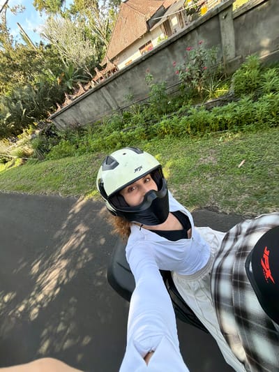 A woman is riding a scooter on a road in Selemadeg, Indonesia. She is wearing a white helmet with black accents, a white zip-up jacket, a black top, white pants, and plaid pants. She is looking at the camera with her eyes wide open and a slight smile. The road is paved and has shadows from trees. There is a concrete wall with decorative elements on the side of the road, and beyond that, lush green vegetation and trees. A building with a tiled roof is visible in the background. The sky is blue with some clouds.